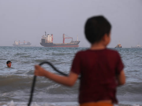 Children play in the water along the shore as a mix of bulk carriers, cargo ships, and service vessels sit offshore in the Strait of Hormuz off Bandar Abbas, Iran, Sunday, April 26, 2026.
