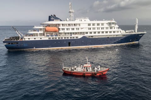 An aerial view of an ambulance boat carrying crew members wearing hazmat suits as they approach the pilot door on the starboard side of the cruise ship MV Hondius, while stationary off the port of Praia, the capital of Cape Verde, on May 5, 2026.