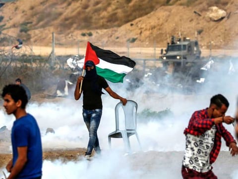 A masked Palestinian protester walks carrying a Palestinian flag and a plastic chair through tear gas fumes amidst clashes with Israeli forces across the barbed wire fence during a protest in the central Gaza Strip.