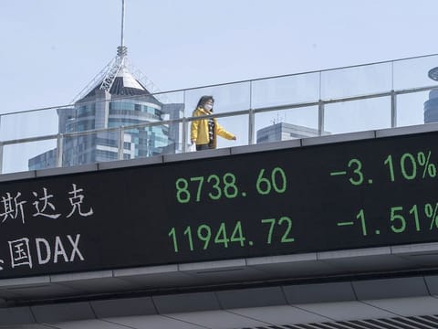 A pedestrian wearing a protective mask walks along an elevated walkway as an electronic ticker displays stock figures in Pudong's Lujiazui Financial District in Shanghai, China, on Friday, March 6, 2020. While infections in the rest of the world accelerate, the coronavirus epidemic is showing signs of easing at its center -- China -- with new cases slowing dramatically and recoveries gathering pace.