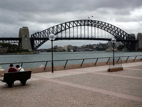 People sit on a bench at the mostly deserted promenade of the Sydney Opera House overlooking the Sydney Harbour Bridge, in the wake of New South Wales implementing measures to prevent the spread of COVID-19 (File)