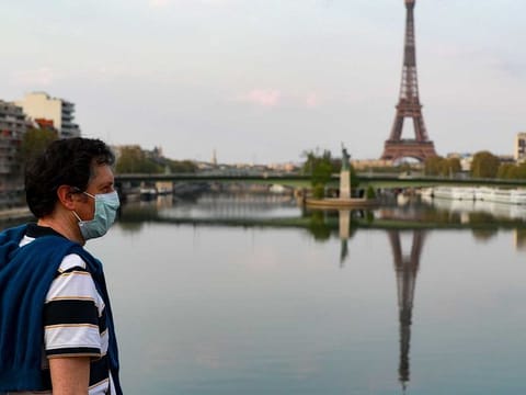 A man wearing a protective facemask looks at the reflection of the Eiffeil Tower on the Seine river, in Paris
