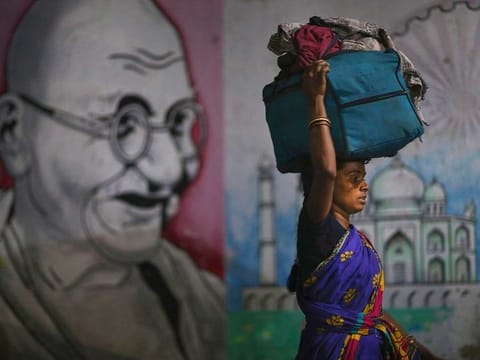 A migrant woman carries her belongings on her head as she walks past a wall painting of Mahatma Gandhi to board a train to her home state, at Hyderabad Railway Station in Hyderabad, India
