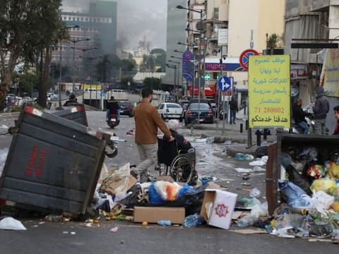 A man pushes an elderly person on a wheelchair near garbage bins placed by demonstrators to block a road, during a protest against the fall in Lebanese pound currency and mounting economic hardships, in Beirut, Lebanon March 16, 2021.
