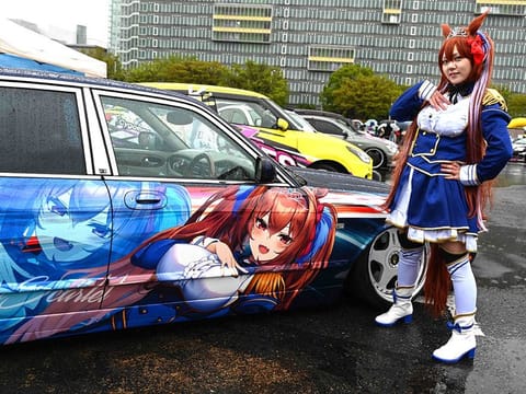 A woman dressed up as anime character Daiwa Scarlet posing next to a car owned by Yosuke Takahata.