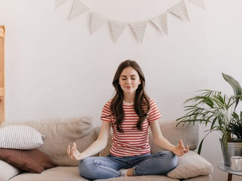 Brunette girl in striped T-shirt is meditating while sitting on sofa in living room.