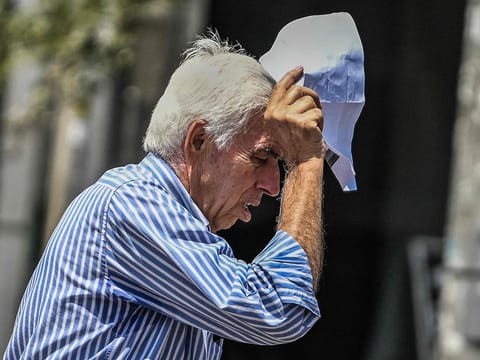 An elderly man holds a piece of paper over his head to protect himself from the sun in Athens.