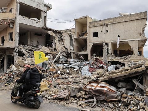 Men ride a scooter with a Hezbollah flag past destroyed buildings as people make their way back to their homes in the south of Lebanon after a ceasefire between Israel and Hezbollah took effect, on November 27, 2024, in the southern town of Qana.