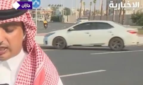 The TV correspondent is giving his report as a car is seen moving in the wrong direction of the road.