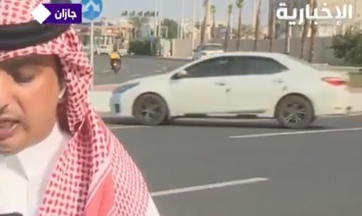 The TV correspondent is giving his report as a car is seen moving in the wrong direction of the road.