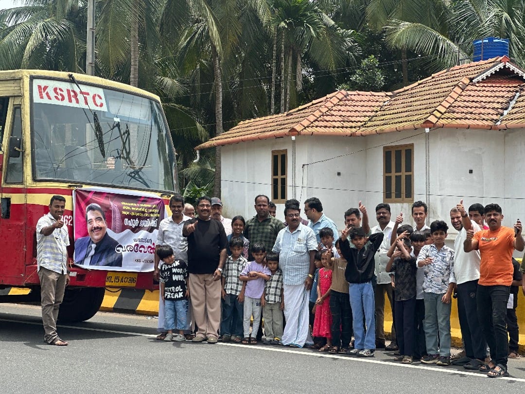 Gafoor, wearing a black t-shirt and light sandy beige pants, smiles and gives a thumbs-up alongside well-wishers by the KSRTC bus bearing a warm welcome banner.