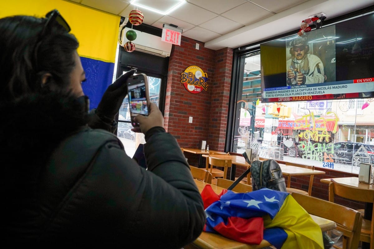 A woman takes a photograph of the television in El Budare Café after President Donald Trump posted a photo of Venezuelan leader Nicolás Maduro aboard the USS Iwo Jima heading for New York, where he faced charges in Manhattan federal court, on January 3, 2026 in the Queens borough of New York City.