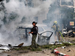 An Israeli policeman looks on as behind a firefighter extinguishes a blaze in a vehicle following a projectile impact in southern Tel Aviv.