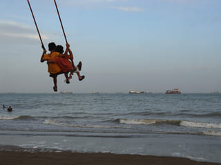 Two children swing together as a mix of bulk carriers, cargo ships, and service vessels line the horizon in the Strait of Hormuz off Bandar Abbas, Iran.