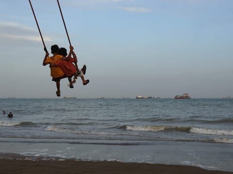 Two children swing together as a mix of bulk carriers, cargo ships, and service vessels line the horizon in the Strait of Hormuz off Bandar Abbas, Iran.