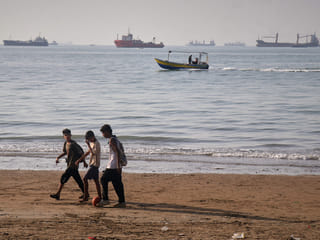 Children walk along the shore with a ball as a small motorboat passes offshore and bulk carriers, cargo ships, and service vessels line the horizon in the Strait of Hormuz off Bandar Abbas, Iran. 