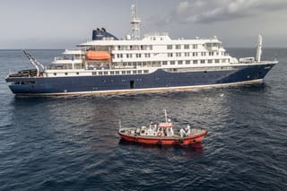 An aerial view of an ambulance boat carrying crew members wearing hazmat suits as they approach the pilot door on the starboard side of the cruise ship MV Hondius, while stationary off the port of Praia, the capital of Cape Verde, on May 5, 2026.