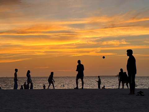 Manjula S. Dadallage captures a vibrant sunset scene at a Dubai beach, where silhouettes of people playing volleyball and enjoying the evening create a striking contrast against the glowing sky and calm shoreline.
