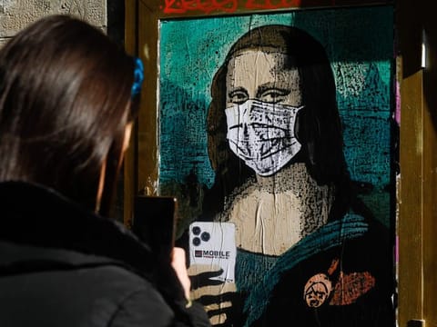 A woman takes a picture with her mobile phone of a poster by Italian urban artist Salvatore Benintende aka "TVBOY" depecting Leonardo da Vinci's Mona Lisa  wearing a protective facemask and holding a mobile phone reading "Mobile World Virus" in a street of Barcelona on February 18, 2020, a week after the World Mobile Congress was cancelled due to fears stemming from the coronavirus that sparked an exodus of industry heavyweights.