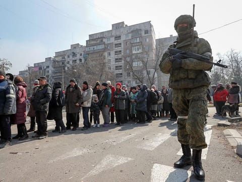 A Russian army soldier stands next to local residents who queue for humanitarian aid delivered during Ukraine-Russia conflict, in the besieged southern port of Mariupol, Ukraine.