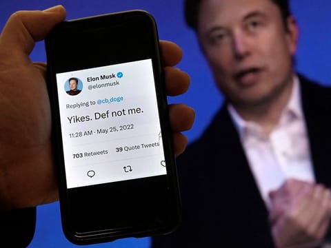 A phone screen displaying a post on Elon Musk's Twitter account stating "Yikes. Def not me" with an allegedly fake video of him promoting a new cryptocurrency scam shown in the background in Washington, DC.