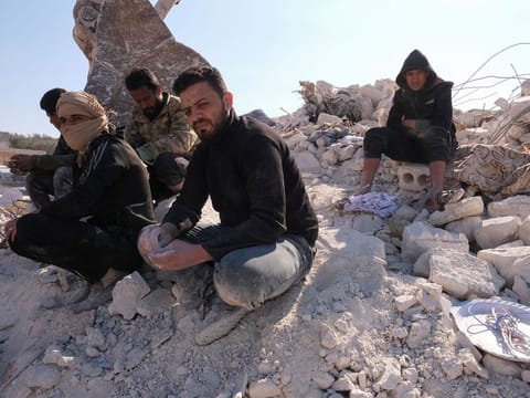 Men with hands covered in dust from sifting through rubble, sit atop ruins in the aftermath of an earthquake in Idlib, Syria, February 14, 2023
