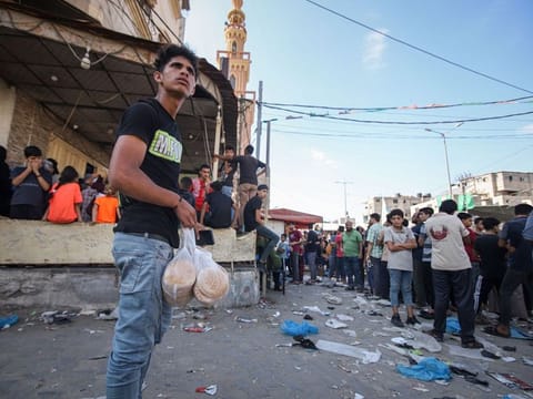 A Palestinian man with bags of fresh bread near a queue of people waiting to collect limited food supplies at a store in Khan Younis, Gaza, on October 16, 2023.  