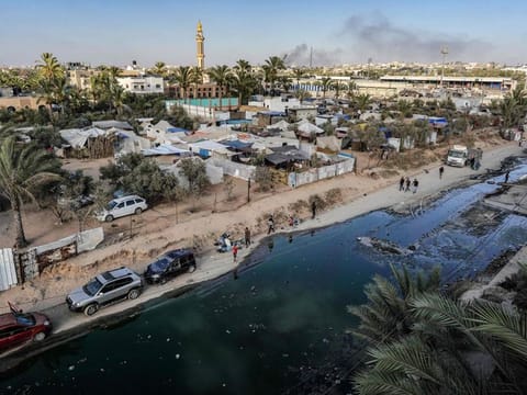 This aerial view shows tents used as temporary shelters for displaced Palestinians along a street covered with stagnant wastewater in Deir Al Balah in the central Gaza Strip on July 19, 2024, as smoke rises from Israeli bombardment in the nearby Nuseirat.  