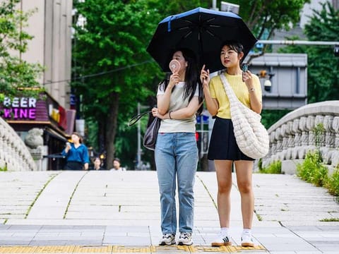 Pedestrians use an umbrella to shield from the sun and a portable fan as they wait to cross a road in Seoul on August 15, 2024, during a prolonged heatwave which has gripped much of the country. 